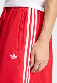 Pantalones cortos deportivos rojos hechos de tela lisa, con rayas blancas en los laterales y un logotipo bordado en el dobladillo, con un bolsillo lateral.