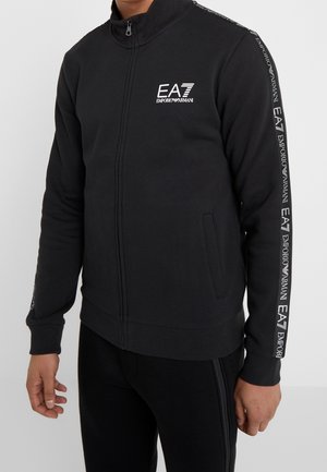 Homme portant une veste noire zippée avec le logo EA7 Emporio Armani sur la poitrine et un ruban de marque le long des manches, associée à un pantalon noir.