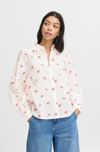 Camisă albă, cu nasturi, cu mici modele de frunze roșii, mâneci lungi, cu mansete și o croială lejeră, asociată cu blugi deschiși la culoare cu talie înaltă.