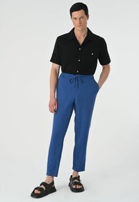 Blauwe, lichte, taps toelopende broek met een elastische tailleband en trekkoord; gecombineerd met een zwart shirt met korte mouwen en zwarte sandalen.