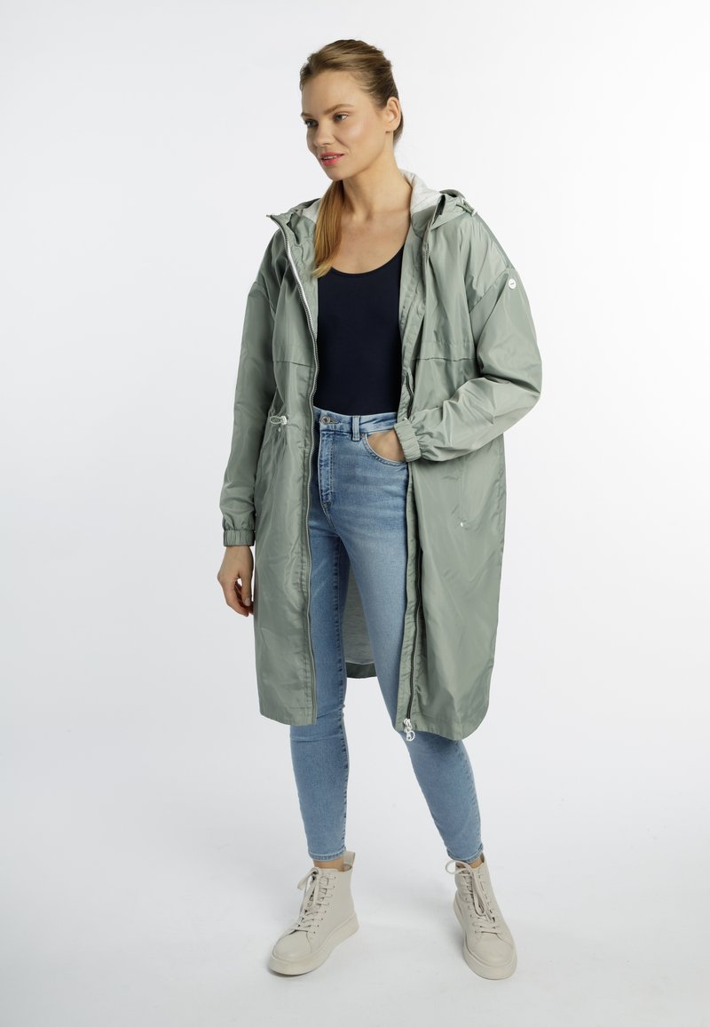 Hellgrüner, langer Regenmantel mit Kapuze, Frontreißverschluss und elastischen Bündchen, kombiniert mit blauen Skinny-Jeans und beigen Sneakers.