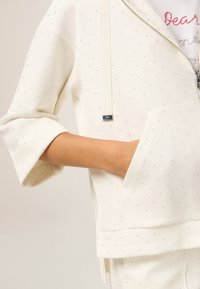Giacca bianca con zip e maniche corte, realizzata in tessuto morbido, con accenti argentati sparsi e dettaglio della tasca anteriore.