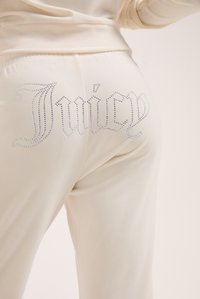 Kremkrāsas sporta bikses ar gludu tekstūru, kas rotātas ar kristālu akcentiem, kas uz aizmugures dekoratīvajā rakstā veido vārdu "Juicy".