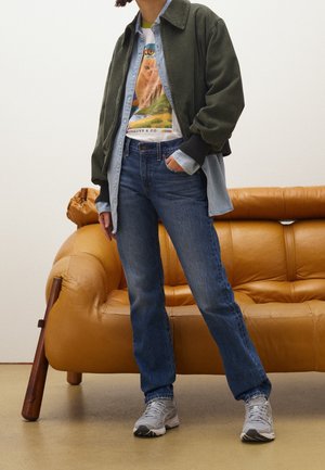 Person står foran en beige sofa iført en grøn jakke, denimskjorte, grafisk kat-T-shirt, blå jeans og grå sneakers.
