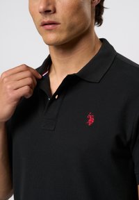 Zwarte katoenen polo met een kraag met drie knopen, voorzien van een rood geborduurd logo en een gestreepte accent aan de binnenkant van de kraag. Close-up weergave.