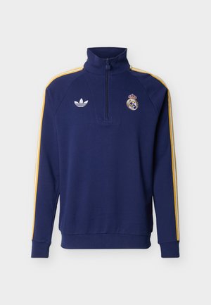 Sudadera azul marino con cremallera hasta el pecho, con franjas amarillas en los hombros, logo de Adidas en el pecho izquierdo y escudo del Real Madrid en el pecho derecho.