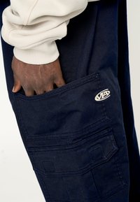 Pantalones cargo azul marino con un gran bolsillo lateral, detalles de costura y un parche de logo circular. La tela parece duradera con una textura suave.