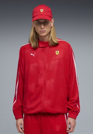 Persona con capelli biondi lunghi che indossa una tuta Puma Ferrari rossa e un cappellino Ferrari rosso coordinato su sfondo grigio.