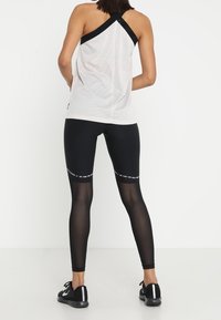 Vrouw in een witte mesh haltertop en zwarte leggings met doorzichtige kuitpanelen en zwarte sportschoenen, staande tegen een witte achtergrond.