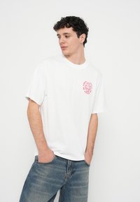 Camiseta branca de algodão com mangas curtas, apresentando um desenho gráfico vermelho no peito esquerdo e um ajuste descontraído, combinada com jeans azuis.