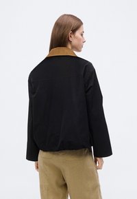 Γυναίκα με ίσια καστανά μαλλιά φοράει μαύρο σακάκι με καφέ γιακά και μπεζ παντελόνι, με την πλάτη της στραμμένη προς ένα απλό ανοιχτό γκρι φόντο.