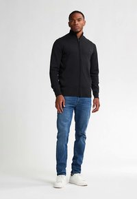 Zwarte trui met ritssluiting en geribbelde textuur, gecombineerd met blauwe jeans en witte sneakers. Model staat recht vooruit tegen een lichte achtergrond.