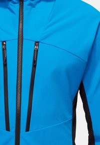 Niebieska kurtka typu softshell o gładkiej fakturze, z dwiema kieszeniami na zamek na piersiach oraz czarnymi akcentami po bokach i przy zamku.