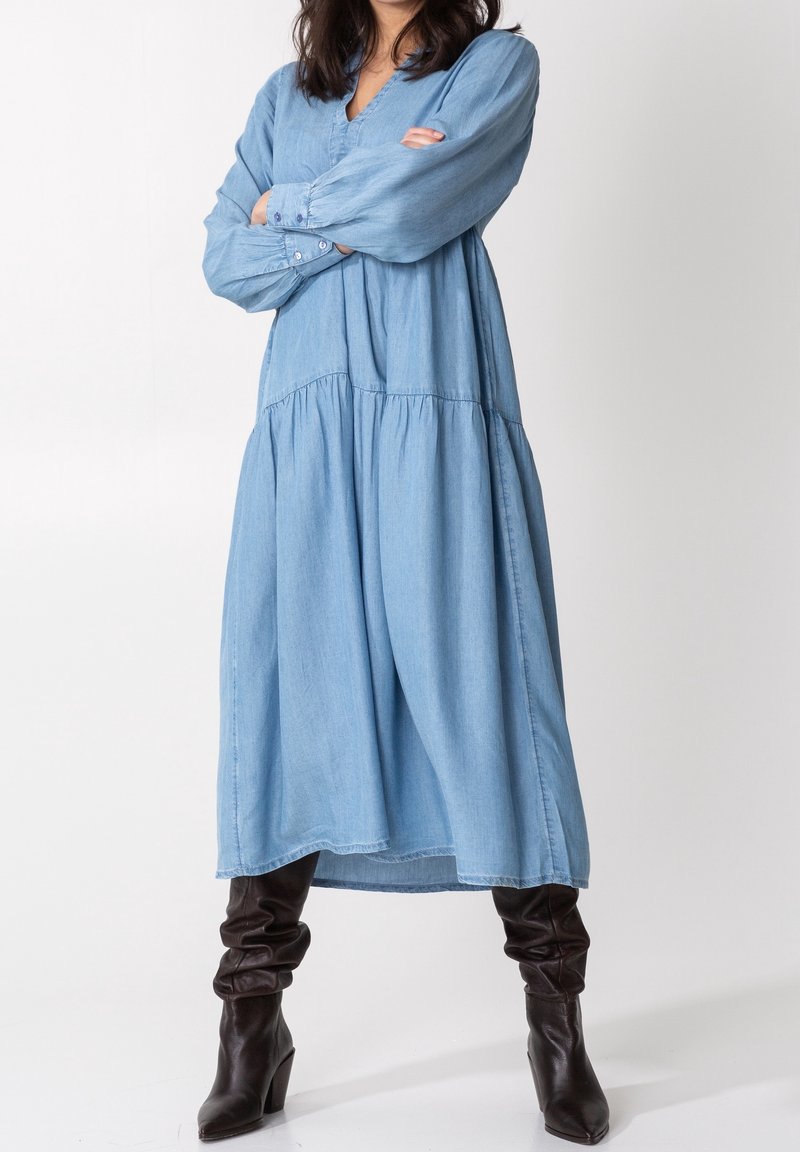 Kobieta ubrana w długą, luźną, jasnoniebieską sukienkę z długimi rękawami i ciemnobrązowe, luźne buty na obcasie, stojąca ze skrzyżowanymi ramionami.