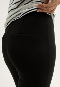 Black fitted trousers with a smooth texture and a single back pocket. The waistband is high-rise, and a striped shirt is partially visible above.