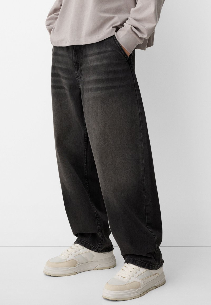 Črne denim hlače s sproščenim krojem, ki vključujejo stranske žepke in subtilno zbledelost. Ujemajo se s svetlo bež supergami, ki se ponašajo z gladko teksturo.