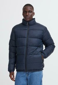 Marineblå pufferjakke med høj krave, quiltet design og lynlås foran. Har sidelommer og elastiske manchetter, der giver isoleret varme.
