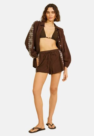 Vrouw die een bruin bikinitopje draagt, bijpassende losse blouse met knopen en bloemrijke borduursels op de mouwen, shorts met trekkoord en zwarte slippers, staand.