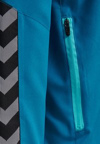 Blauwe sportkleding met een textuur zwart zijpaneel dat een wit chevronpatroon heeft. Inclusief een zak met rits met een turkooizen rits.