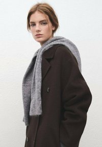 Bruine wollen jas met een dubbelbreasted ontwerp en zwarte knopen, gecombineerd met een lichtgrijze wollen sjaal, die zachte texturen laat zien.