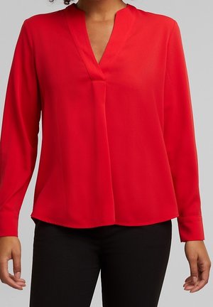 Kvinde iført en rød V-hals bluse med lange ærmer og sorte bukser, stående mod en ensfarvet grå baggrund.