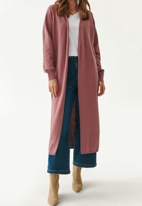 Žena v dlouhém mauve kardiganu, bílém tričku s výstřihem do V, modrých širokých džínách a béžových špičatých botách, stojící na jednobarevném pozadí.