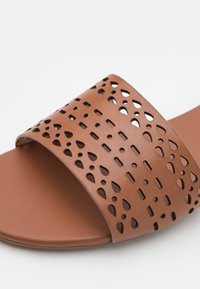 Sandale à glissière marron avec une large sangle présentant des motifs de découpes rectangulaires et en forme de goutte d'eau.