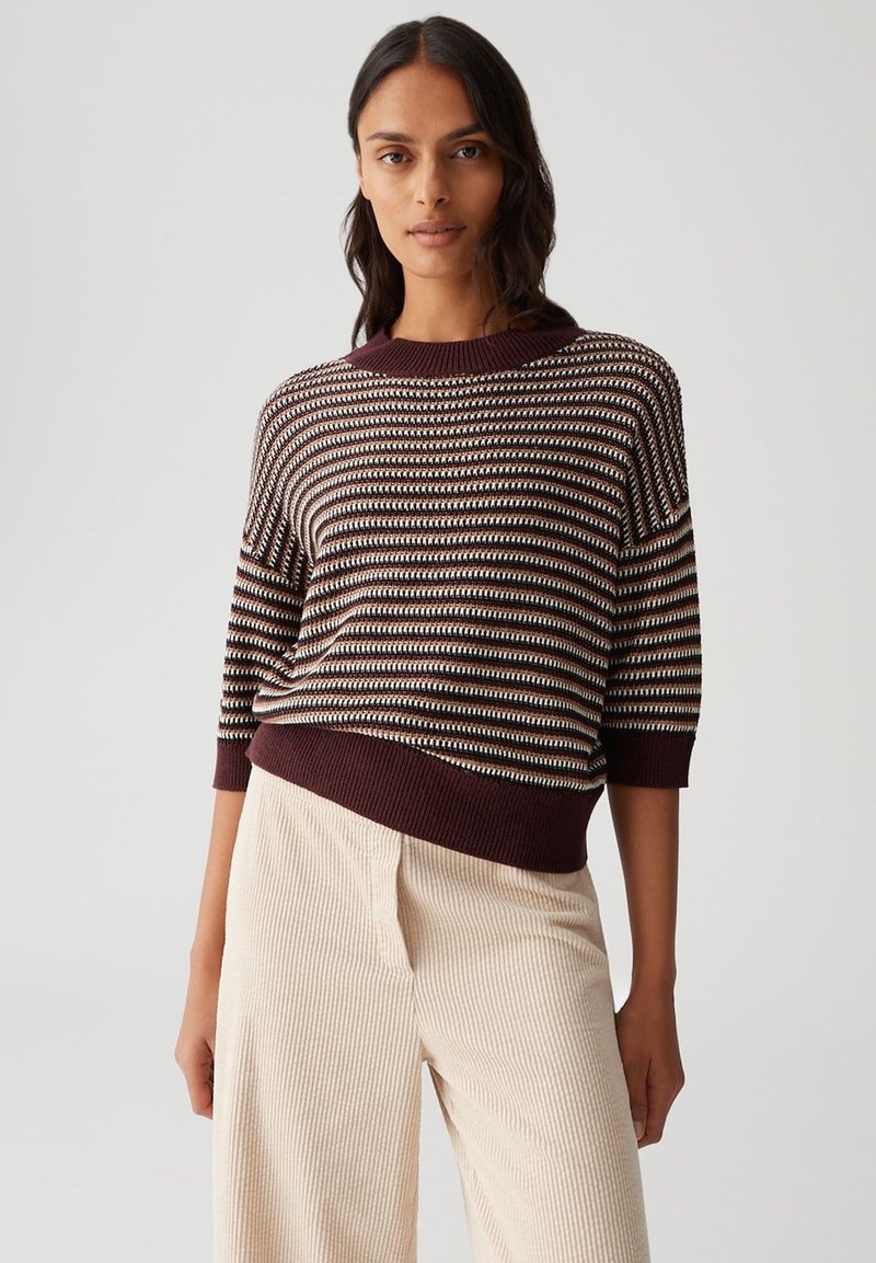 Woman wearing a brown, black, and white striped sweater with three-quarter sleeves and high-waisted beige corduroy pants against a plain background.