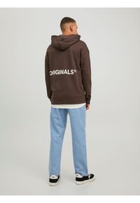 Hoodie marron avec un grand logo blanc "ORIGINALS™" dans le dos, associé à un pantalon en denim bleu clair et des baskets noires.