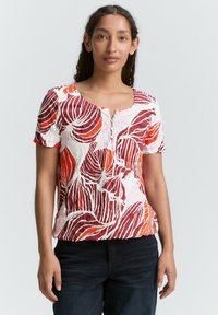 Haut à manches courtes blanc avec des motifs de feuilles rouges et oranges ; il présente un col rond et des boutons décoratifs à l'avant.
