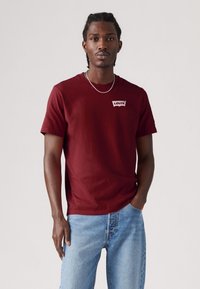 Fonott hajú férfi, bordó Levi's pólót és világoskék farmernadrágot viselve, egy sima fehér háttér előtt áll.