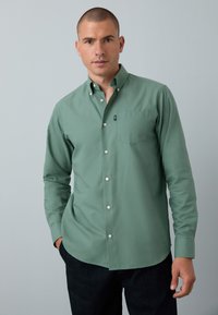 Camicia a maniche lunghe di colore verde chiaro con bottoni e taschino, tessuto liscio e bottoni bianchi. Indossata con le mani nelle tasche contro uno sfondo neutro.