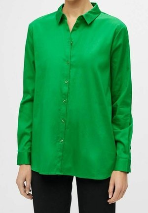 Kvinde iført en langærmet, button-up skarp grøn skjorte med krave, kombineret med sorte bukser, stående mod en ensfarvet baggrund.