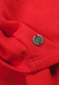 Czerwony materiał o teksturze z tkanym wzorem, z okrągłym metalowym guzikiem z wytłaczanym wzorem, przyszytym czerwonym przeszyciem.