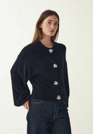 Černý pletený kardigan s žebrovaným lemem, zdobený velkými knoflíky s kovovými detaily a širokými rukávy. Kombinováno s tmavými džínami.