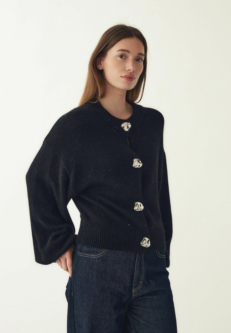 Černý pletený kardigan s žebrovaným lemem, zdobený velkými knoflíky s kovovými detaily a širokými rukávy. Kombinováno s tmavými džínami.