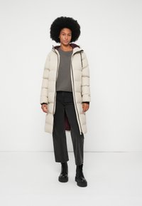 Beige lang puffer jakke med hætte, med lynlås foran, sorte detaljer og en bordeaux indvendig, parret med en grå skjorte og sorte bukser.
