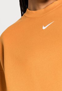Orange sweatshirt i mjukt tyg, med hög krage och en vit Nike-logotyp på bröstet. Slät struktur, avslappnad passform.