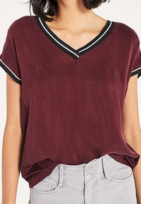 Burgundy short-sleeved top with a V-neck, featuring a black and white striped trim. Smooth texture and relaxed fit. Paired with light gray jeans.