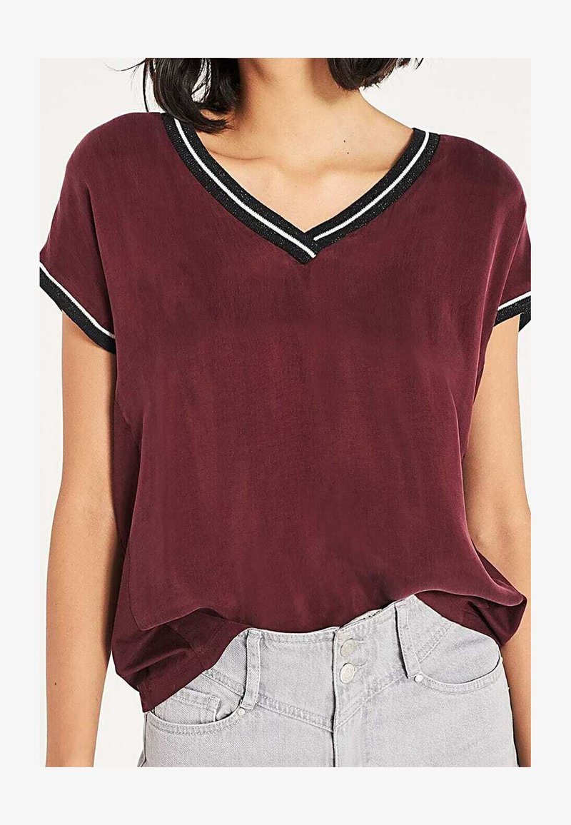 Burgundy short-sleeved top with a V-neck, featuring a black and white striped trim. Smooth texture and relaxed fit. Paired with light gray jeans.
