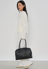 Femme en pull blanc et pantalon tenant un grand sac fourre-tout en cuir noir, debout devant un fond gris clair uni.