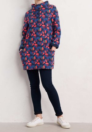 Kobieta w granatowej tunice z czerwonym i zielonym wzorem liści, ciemnych dopasowanych spodniach, białych trampkach, stojąca oparta o jasną ścianę z rękami w kieszeniach.