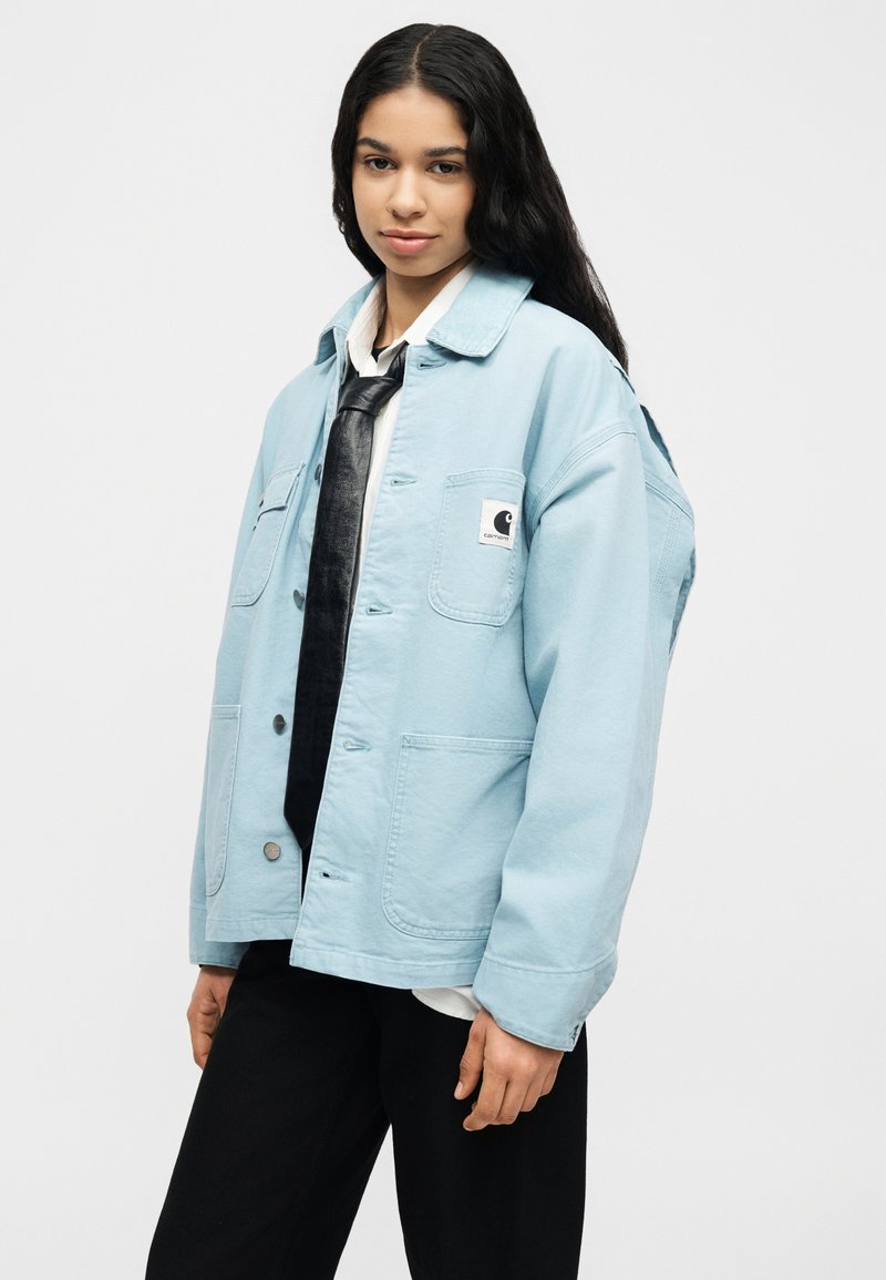 Jonge vrouw met lang donker haar, gekleed in een lichtblauw Carhartt-jasje, wit shirt, zwarte stropdas en zwarte broek, tegen een effen achtergrond.