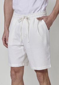 Uomo che indossa pantaloni corti bianchi a coulisse con una texture e una maglietta bianca, con una mano in tasca, su uno sfondo semplice.