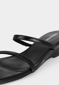 Μαύρο σανδάλι ανοιχτού δακτύλου με δύο λεπτούς ιμάντες πάνω από το πόδι και χαμηλό, χοντρό τακούνι σε ανοιχτό φόντο.