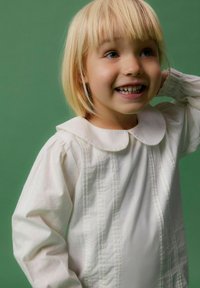 B LJoknlgbetalingjoknlwiuekind met blond haar, gekleed in een witte longsleeve met een Peter Pan-kraag, geposeerd tegen een groene achtergrond.