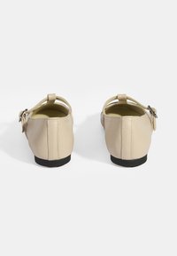 Beige Leder Schuhe von hinten mit runder Spitze, verstellbarem Schnallenriemen und einer niedrigen schwarzen Gummisohle. Glatte Textur, minimalistisches Design.