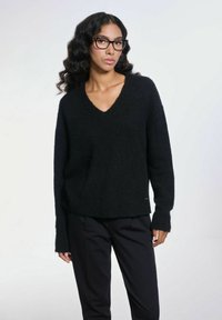 Czarny sweter w serek z miękkiego, teksturowanego materiału, z długimi rękawami i lekko skróconym fasonem. Noszony na czarnych spodniach.