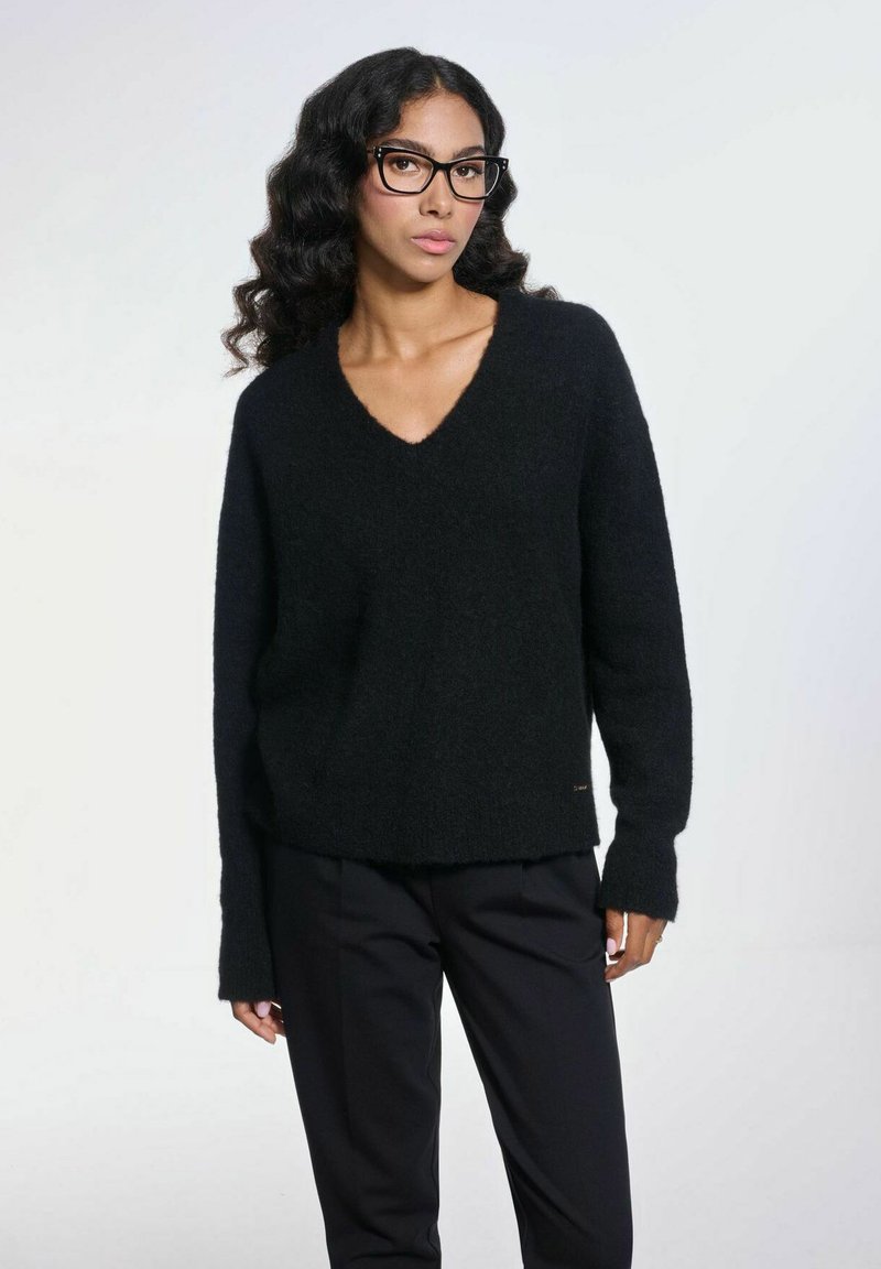 Czarny sweter w serek z miękkiego, teksturowanego materiału, z długimi rękawami i lekko skróconym fasonem. Noszony na czarnych spodniach.