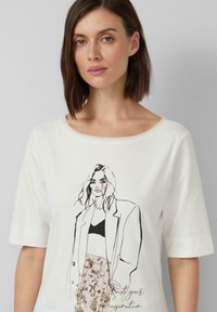 T-shirt blanc à manches courtes avec un croquis en noir et blanc d'une femme portant un blazer, avec une partie inférieure à motif floral et du texte.